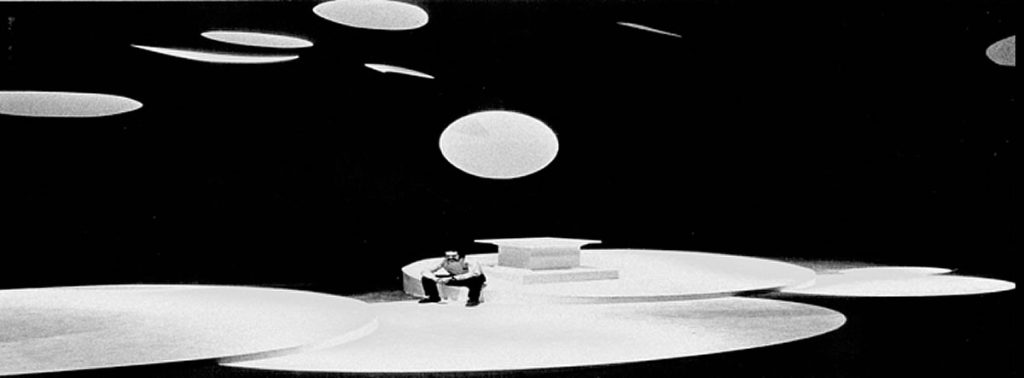 photograph of a modern designed abstract stage with a man sitting in the center on the edge of a rounded raised piece of the stage